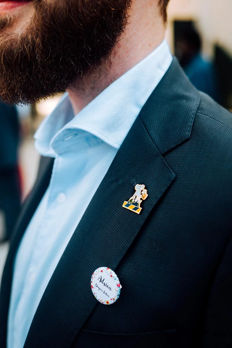 Gros plan d'un homme barbu en costume bleu marine et chemise claire, portant un badge nominatif et un pin's doré.