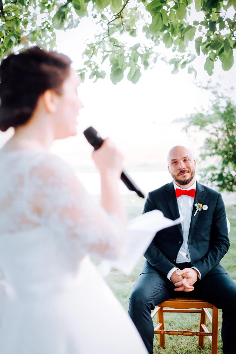 Une mariée prononce son vœu au micro face à son futur époux en costume sombre et nœud papillon rouge.