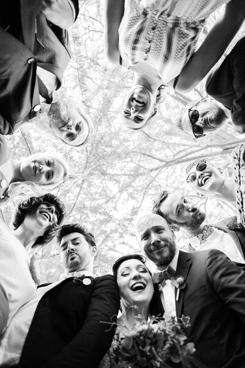 Photo en noir et blanc d'un groupe d'amis souriants entourant les mariés, vue en contre-plongée sous des arbres.