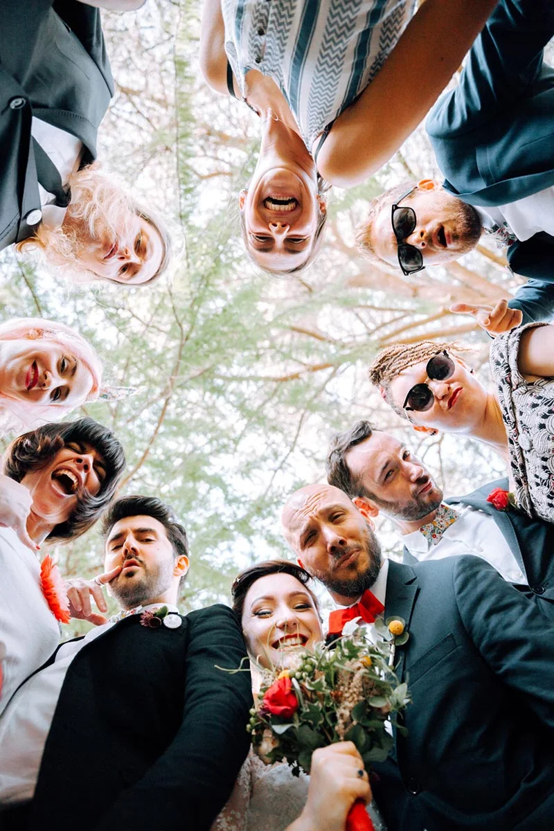 Groupe d'amis et mariés souriants vus en contre-plongée sous des arbres, ambiance festive et naturelle.