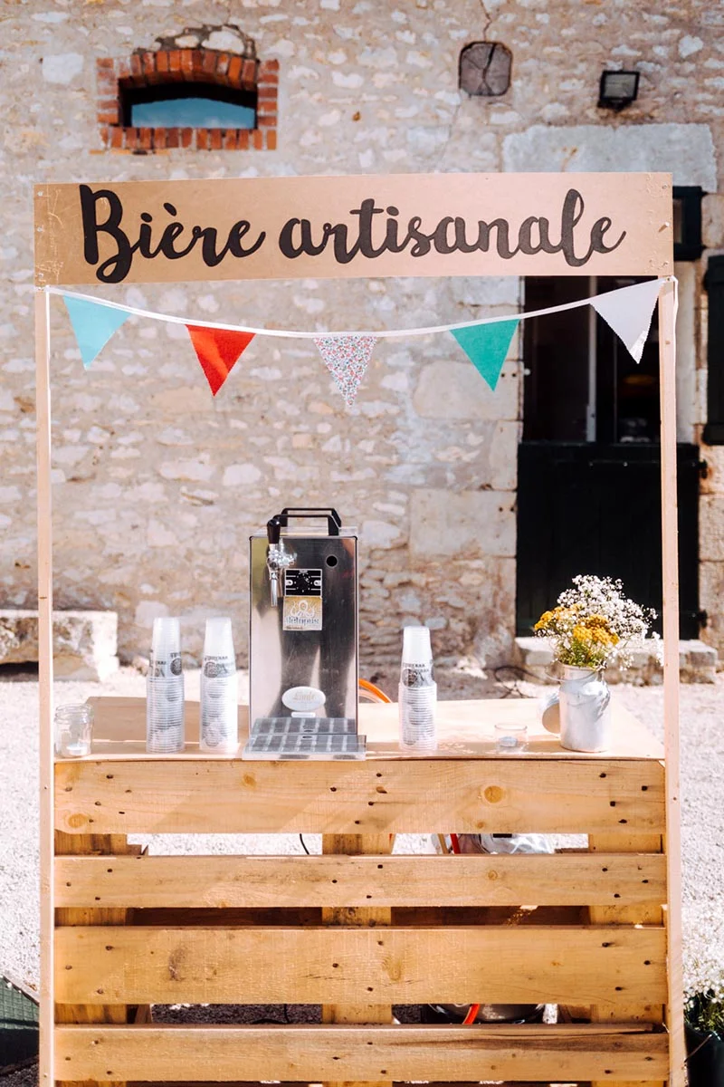 Bar à bière artisanale en bois avec guirlandes colorées devant un mur en pierre pour le mariage de Mel & Ben.