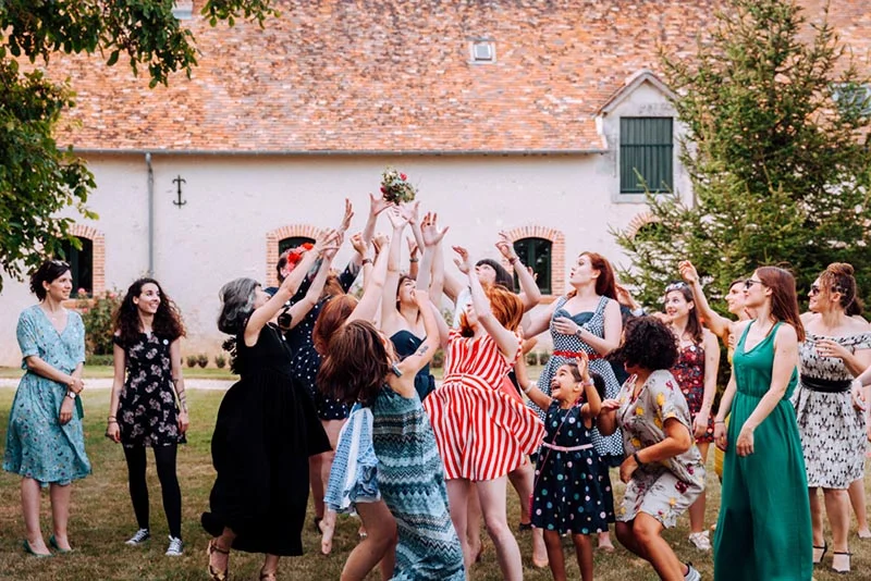 Un groupe de femmes en robes colorées lance le bouquet de la mariée dans le jardin d'une bâtisse rustique.