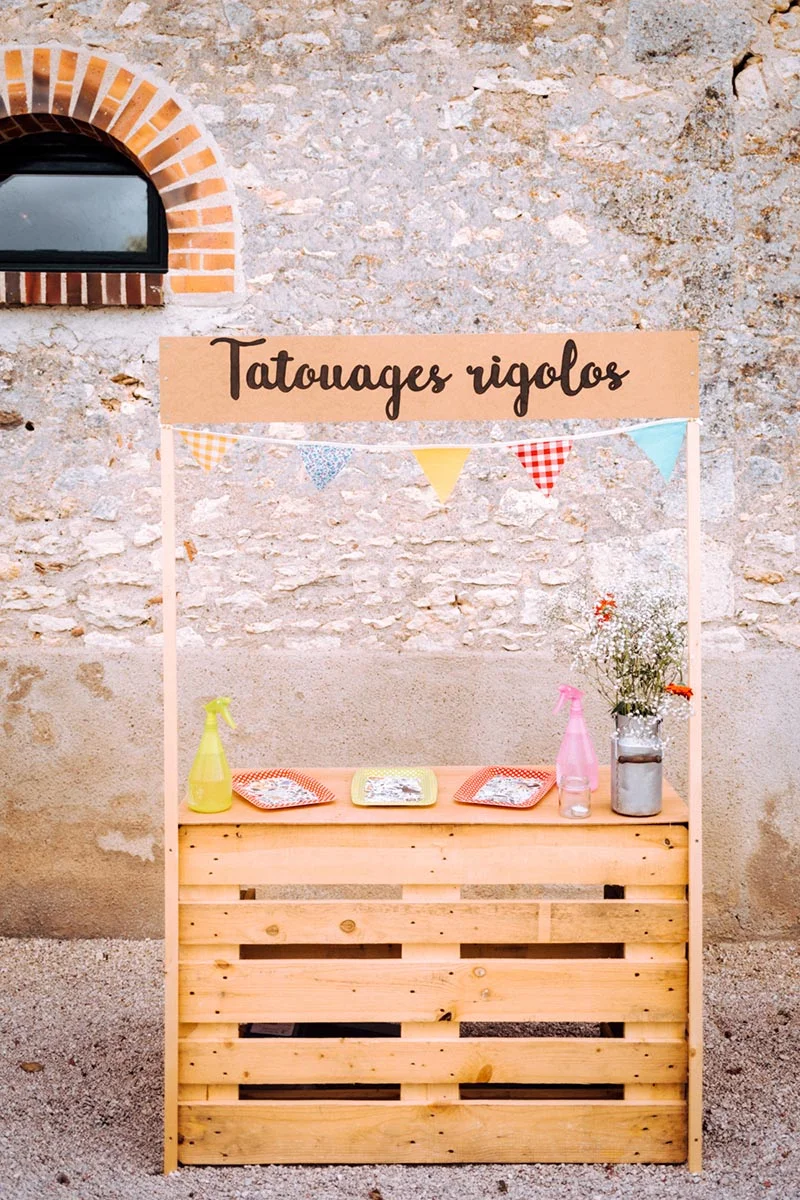 Stand de tatouages rigolos en bois clair avec fanions colorés devant un mur en pierre pour le mariage de Mel & Ben.