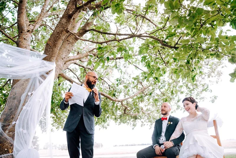 Un officiant s'adresse à Mel et Ben, souriants, lors de leur cérémonie de mariage en plein air sous un grand arbre.