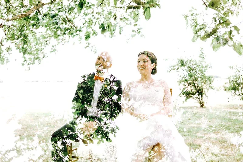 Couple de mariés souriants sous un feuillage lumineux, dans une ambiance romantique et vaporeuse.