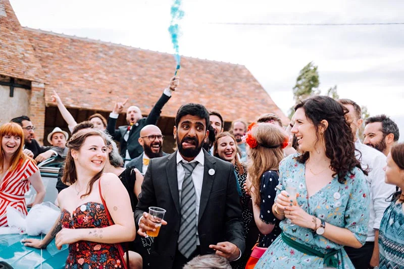 Un groupe d'invités joyeux et colorés célèbre un mariage avec un fumigène bleu devant une grange en briques.