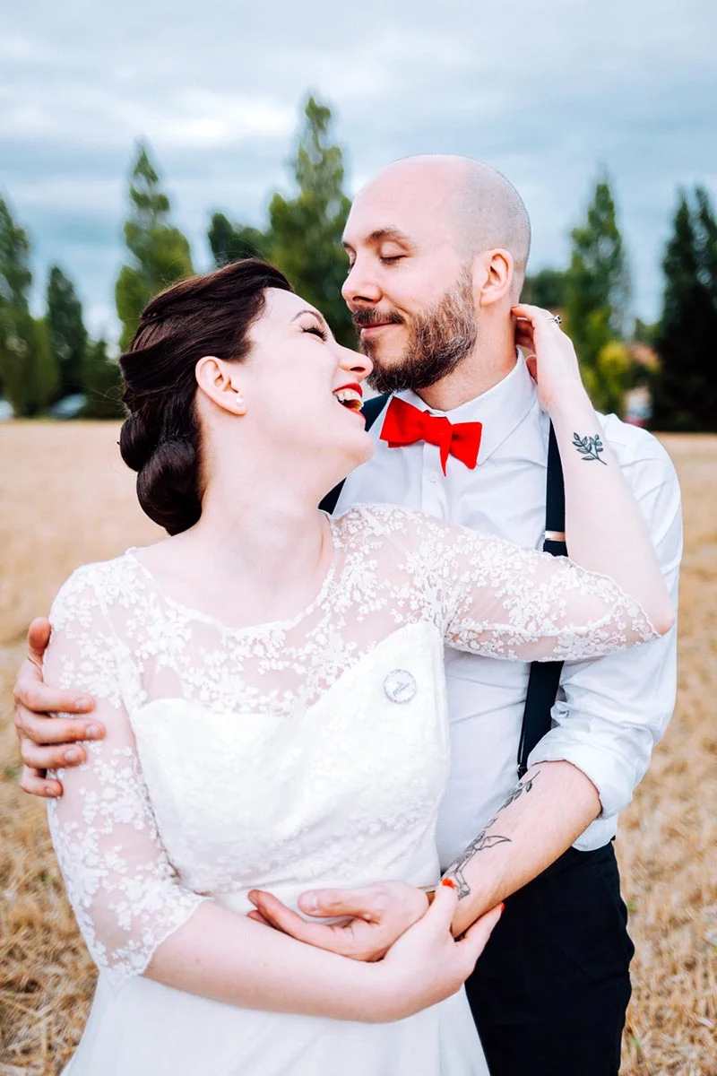 Couple riant tendrement en tenue de mariage, elle en robe de dentelle blanche et lui avec un nœud papillon rouge.