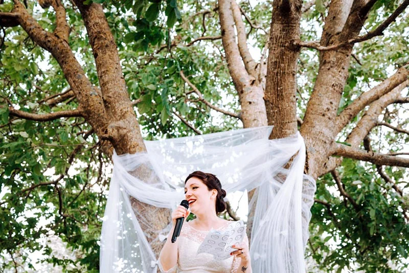 Une mariée souriante prononce un discours au micro sous un arbre décoré d'un voile blanc vaporeux.