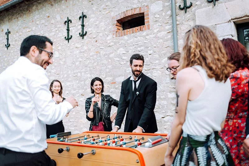 Des invités sourient et s'amusent autour d'un baby-foot lors du mariage de Mel et Ben, devant un mur en pierre.