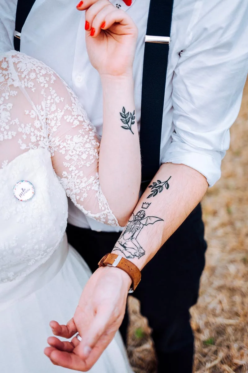 Gros plan sur les bras entrelacés de Mel et Ben, mettant en avant leurs tatouages et leurs tenues de mariage blanches et noires.