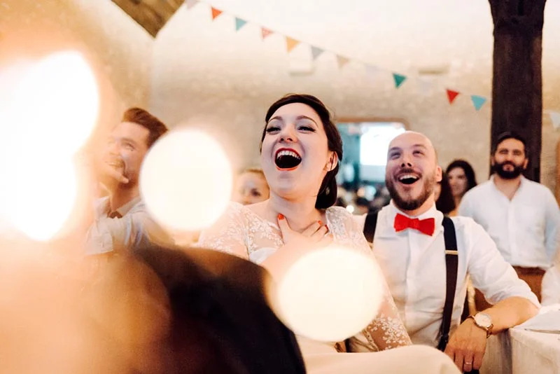 Jeunes mariés riant aux éclats lors de leur réception, ambiance festive et lumineuse avec des guirlandes colorées.