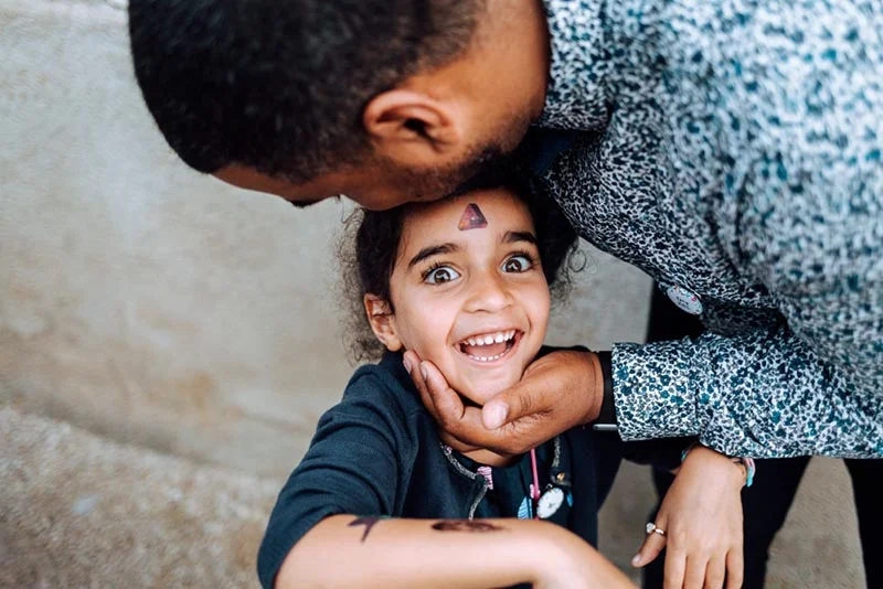Un homme embrasse tendrement le front d'une petite fille souriante, dans une ambiance chaleureuse et intime.