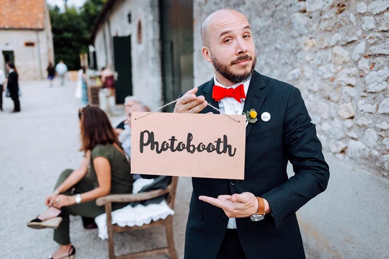Homme en costume noir et nœud papillon rouge tenant un panneau Photobooth lors du mariage de Mel & Ben.