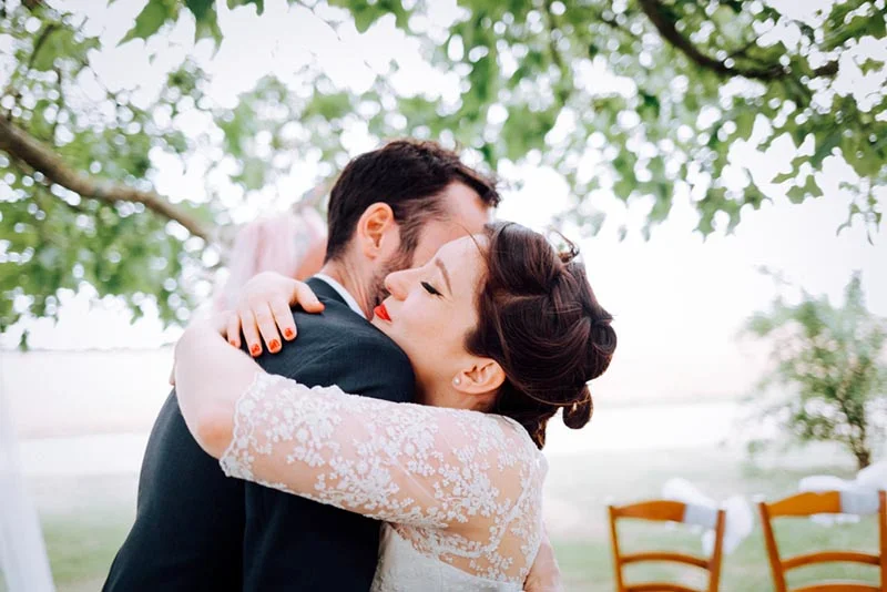 Jeune couple de mariés s'enlaçant tendrement sous un arbre, elle en robe de dentelle blanche et lui en costume sombre.