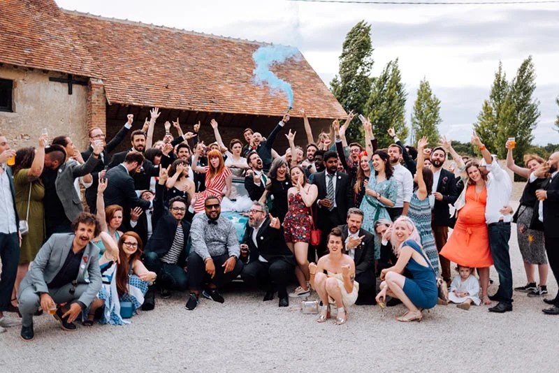Groupe d'invités joyeux célébrant le mariage de Mel et Ben devant une grange, avec un fumigène bleu et une voiture ancienne.
