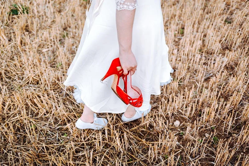 Une mariée en robe blanche tenant des escarpins rouges à la main dans un champ de blé doré.