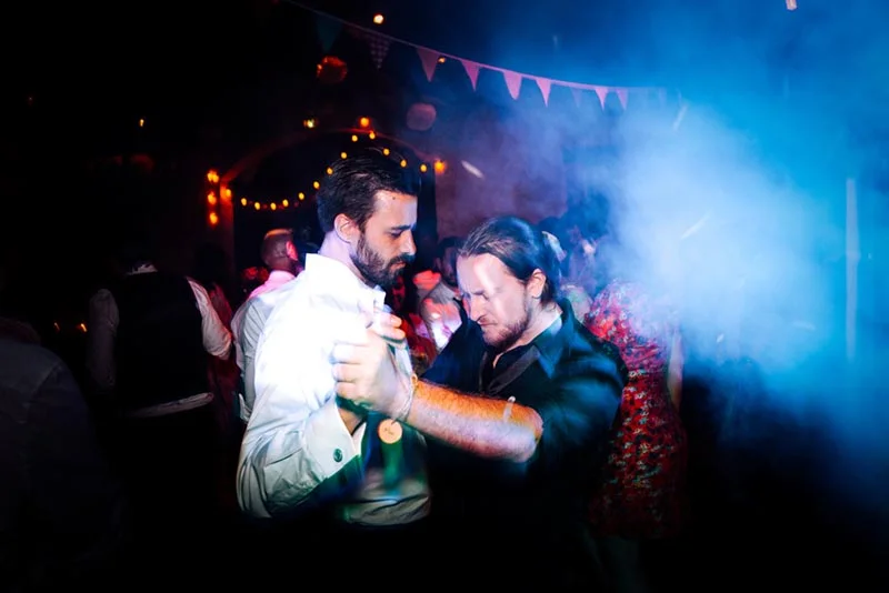 Deux hommes dansent ensemble lors d'une soirée festive sous des lumières bleues et des guirlandes colorées.