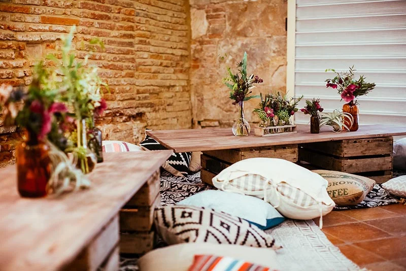Espace lounge bohème avec tables en bois, coussins blancs et bouquets colorés devant un mur de briques anciennes.