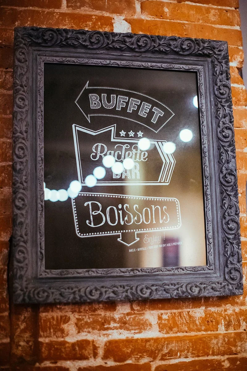 Ardoise encadrée d'un cadre baroque gris sur un mur de briques, indiquant Buffet, Raclette Bar et Boissons.