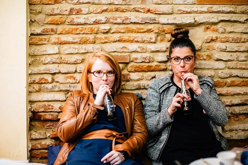 Deux femmes à lunettes, l'une en veste marron et l'autre en veste grise, boivent à la bouteille devant un mur de briques.