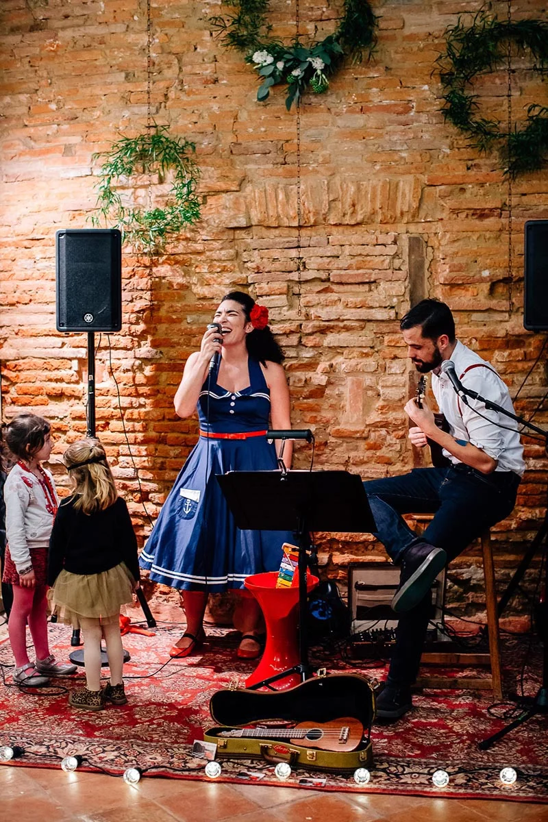 Chanteuse en robe bleue et musicien animent un mariage dans un cadre rustique en briques avec deux petites filles.