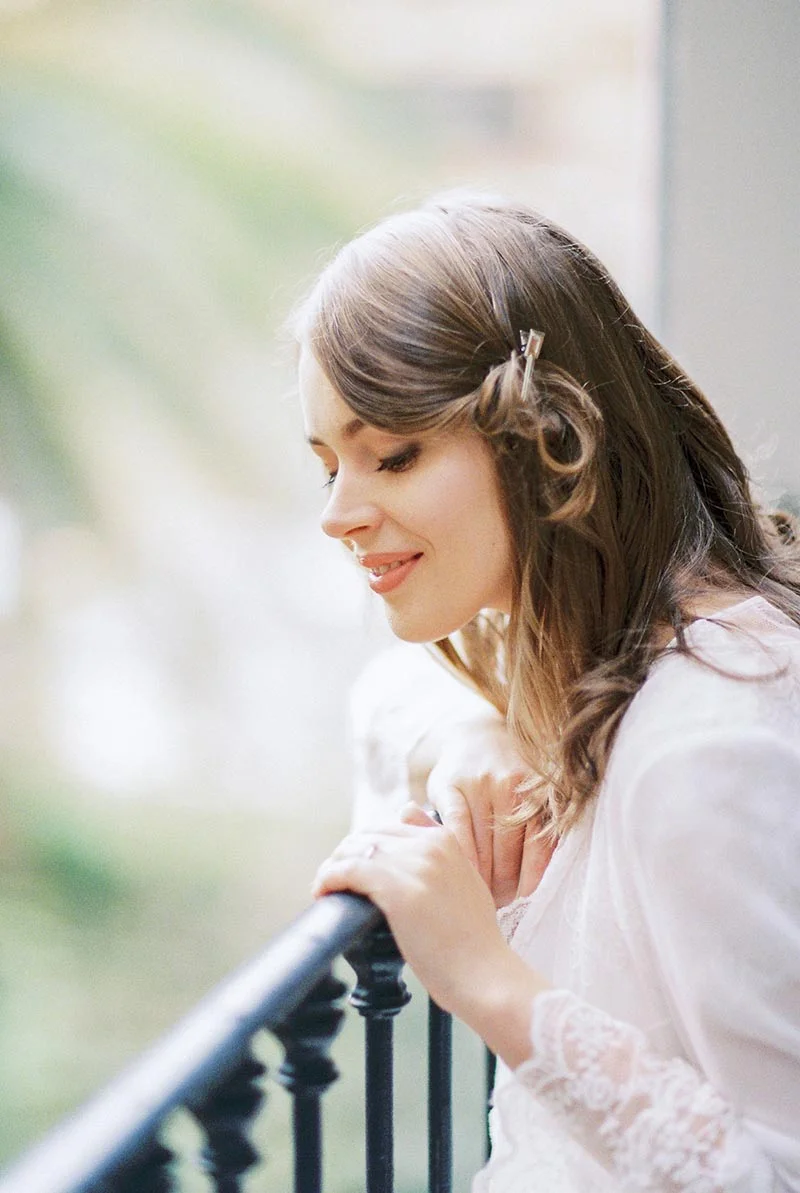 Portrait romantique de Julia, souriante et pensive, vêtue de blanc et appuyée contre un balcon en fer forgé.