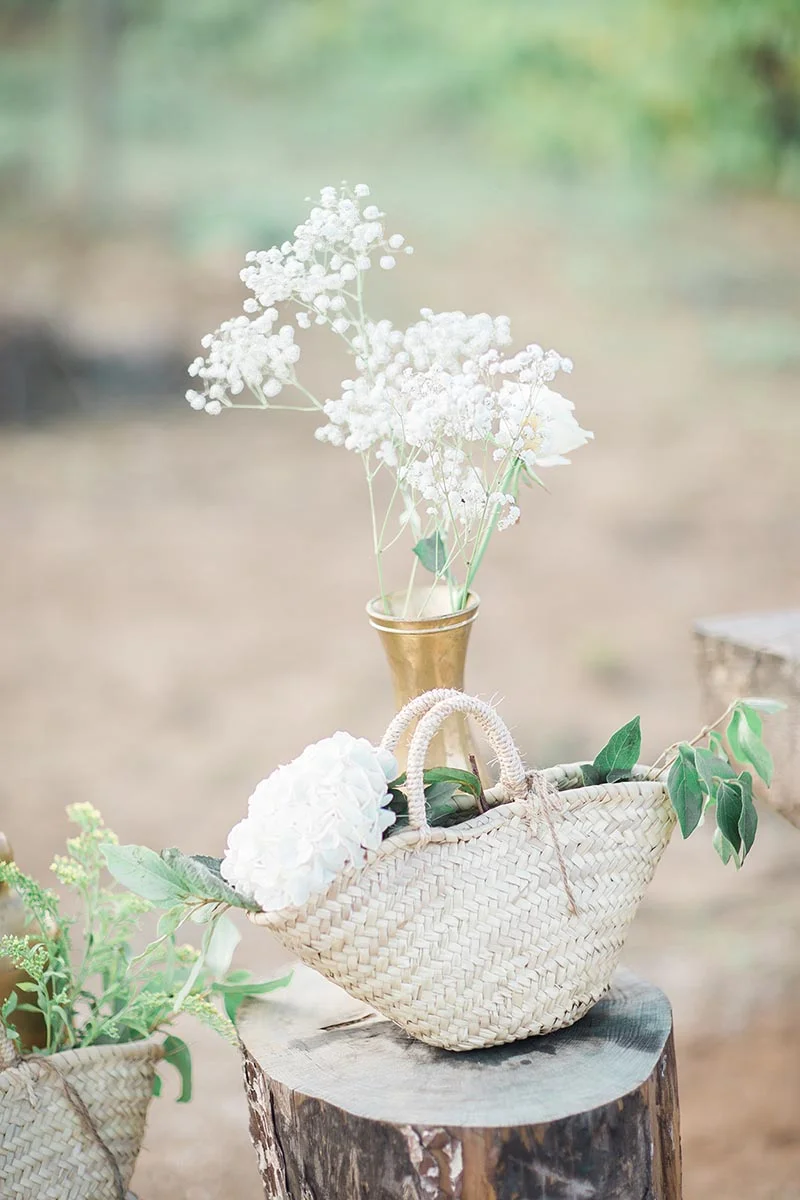 Composition champêtre avec gypsophile dans un vase doré et fleurs blanches dans un panier en osier sur une souche.