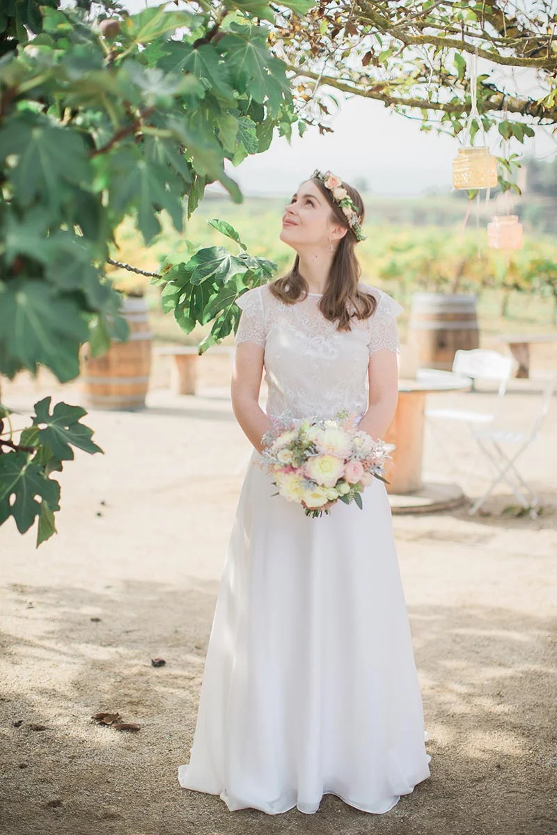 Une mariée en robe blanche avec couronne de fleurs et bouquet, posant sous une treille dans un vignoble lumineux.