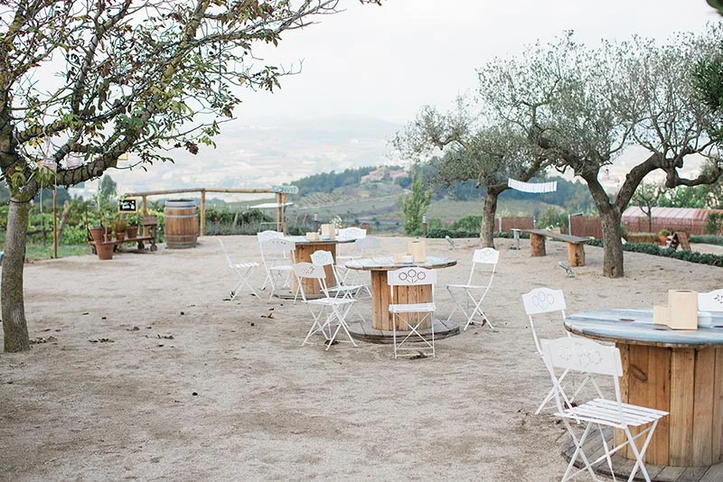 Espace cocktail champêtre avec tables en bois et chaises blanches sous des oliviers, vue sur les collines toscanes.