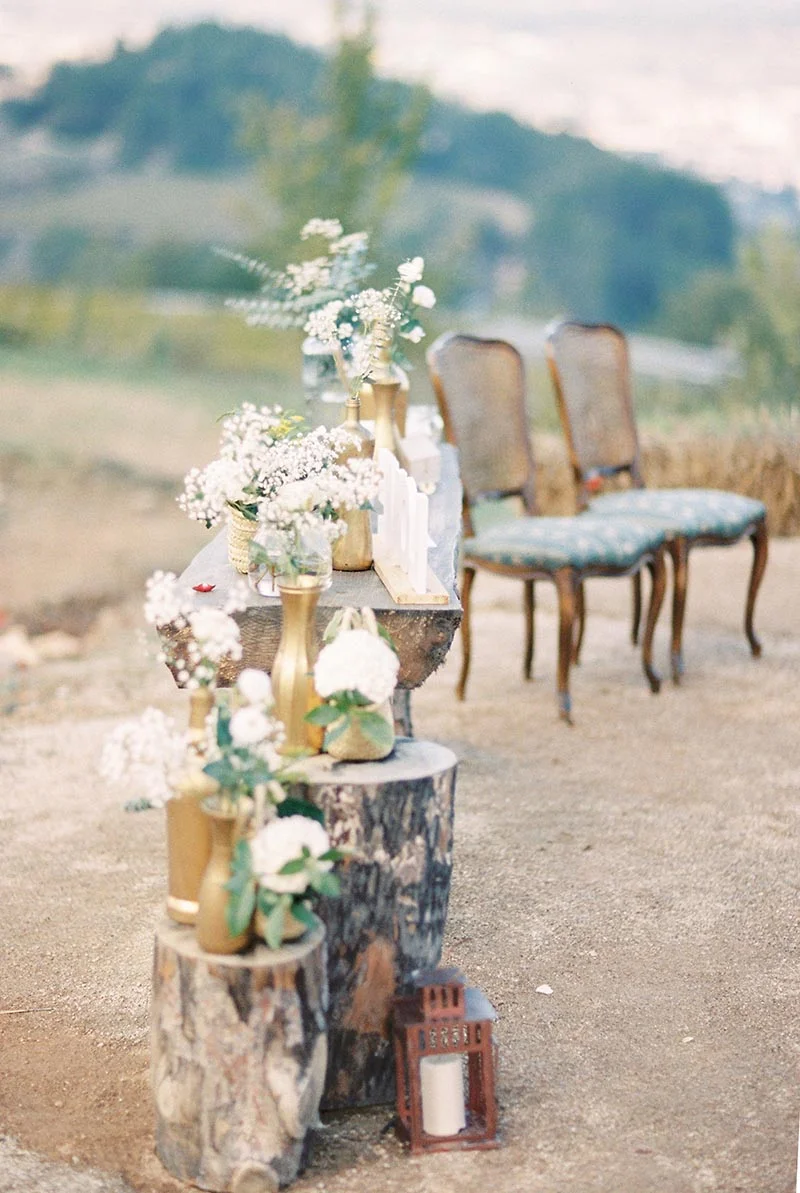 Décoration champêtre avec fleurs blanches et vases dorés sur des rondins de bois, devant deux chaises vintage.