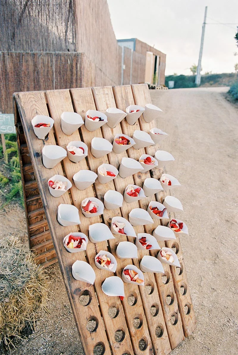 Petits cônes en papier kraft remplis de pétales rouges et blancs disposés sur un support en bois rustique.