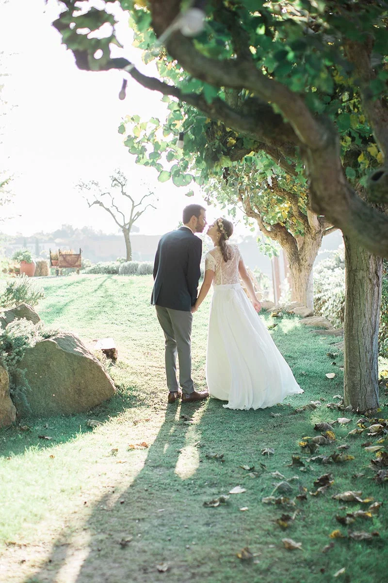 Jeunes mariés s'embrassant sous un arbre feuillu dans un jardin lumineux, ambiance romantique et naturelle.