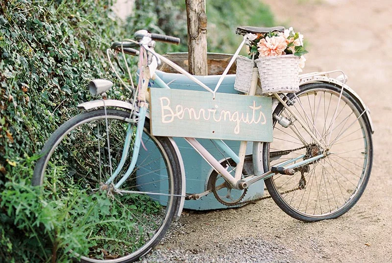 Vélo vintage bleu ciel avec panier de fleurs et panneau Benvinguts pour l'accueil du mariage de Julia et Maxime.