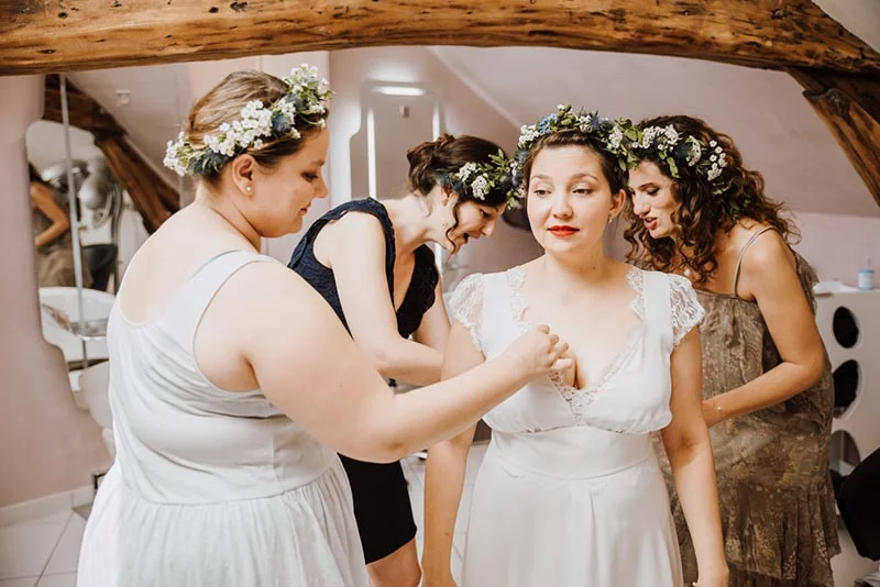 Trois demoiselles d'honneur avec des couronnes de fleurs aident la mariée en robe blanche à s'habiller.