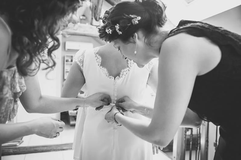 Deux femmes aident une mariée à boutonner sa robe blanche dans une ambiance intime, cliché en noir et blanc.