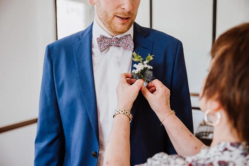 Une femme ajuste la boutonnière fleurie sur le costume bleu d'un homme portant un nœud papillon coloré.