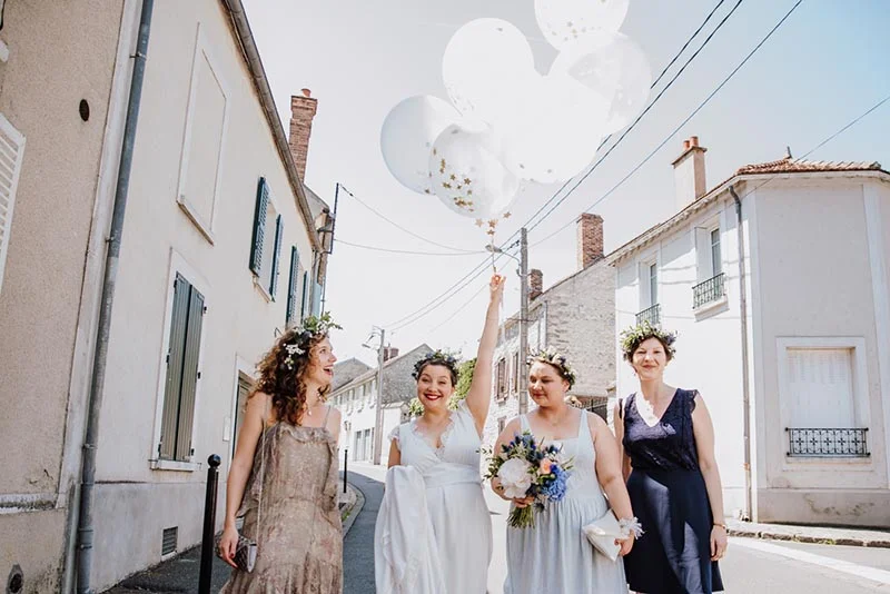 Quatre femmes souriantes en robes claires et couronnes de fleurs marchent dans une rue, tenant des ballons blancs.