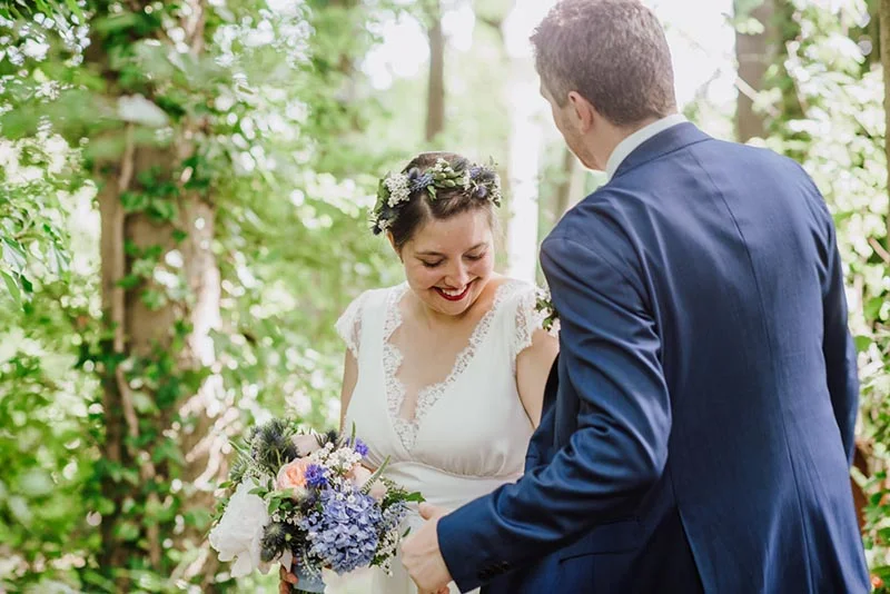 Jeune mariée en robe blanche et couronne de fleurs souriant à son époux en costume bleu dans un cadre naturel verdoyant.