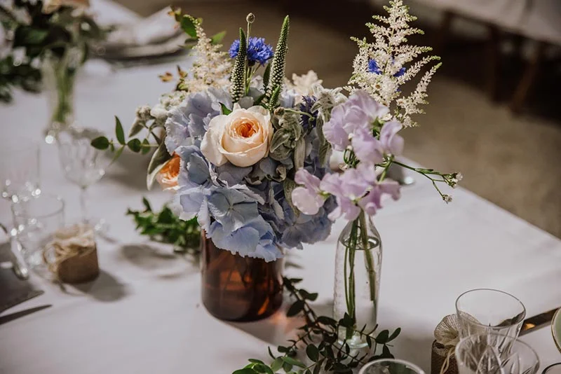 Composition florale romantique aux tons pastel, bleu et blanc, dans des vases sur une table de mariage nappée de blanc.