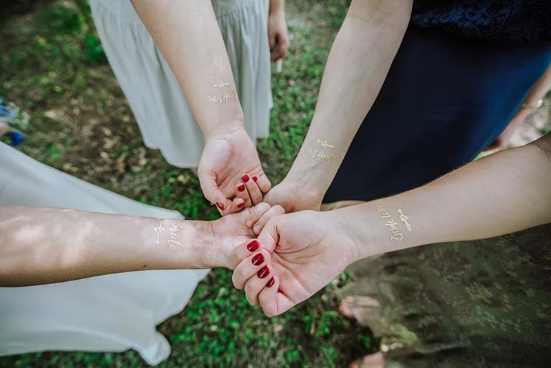 Quatre mains se rejoignent, arborant des tatouages dorés Bride et Bride Tribe sur un fond de nature verdoyante.