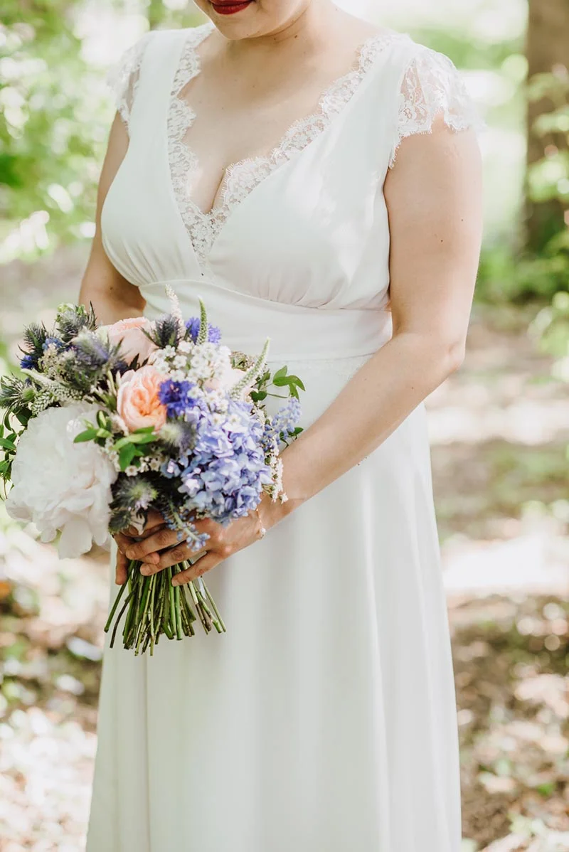 Mariée en robe blanche avec dentelle tenant un bouquet champêtre bleu, blanc et rose dans un cadre naturel.