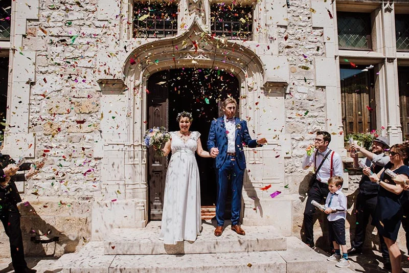 Jeunes mariés sortant d'une église sous une pluie de confettis colorés, entourés de leurs invités.