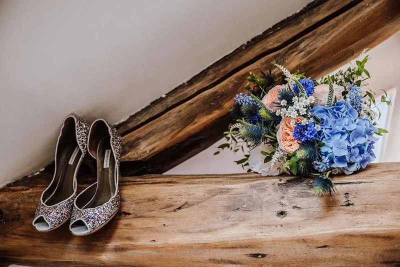 Chaussures à paillettes argentées et bouquet de fleurs bleues et roses posés sur une poutre en bois rustique.