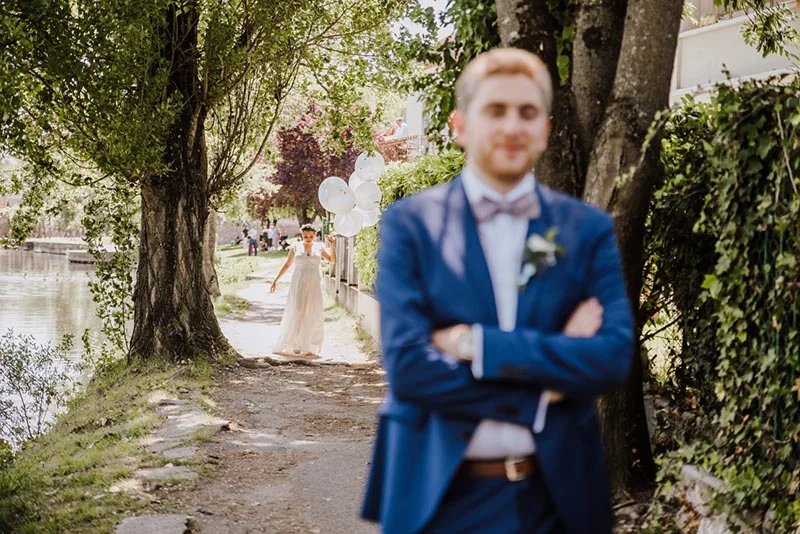 Un marié en costume bleu au premier plan, flou, tandis qu'une mariée en robe blanche arrive avec des ballons blancs.