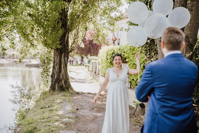 Une mariée souriante en robe blanche tenant des ballons blancs face à son futur époux en costume bleu dans un jardin.