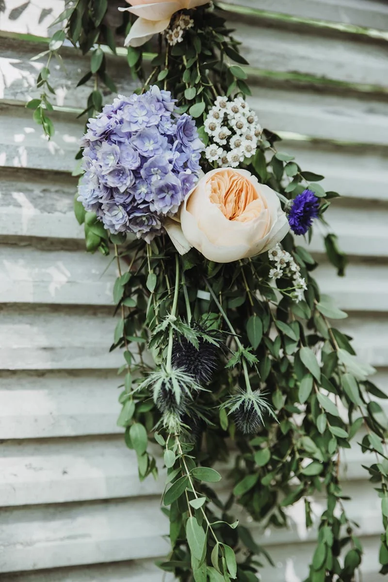 Composition florale romantique avec hortensias mauves, rose pêche et eucalyptus sur un mur blanc.