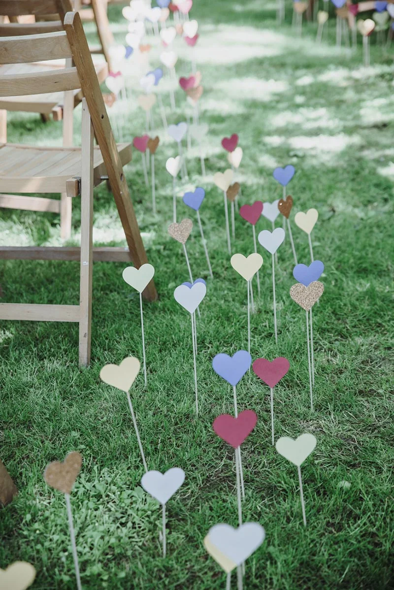 Petits cœurs colorés plantés dans l'herbe le long d'une allée de chaises en bois pour un mariage champêtre.