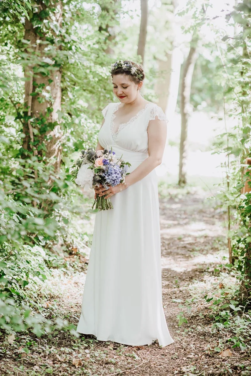 Mariée en robe blanche et couronne de fleurs tenant un bouquet coloré dans un sous-bois lumineux.