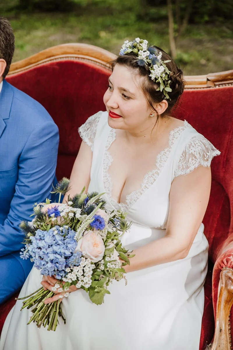 Une mariée souriante en robe blanche avec une couronne de fleurs et un bouquet bleu, assise sur un canapé rouge.