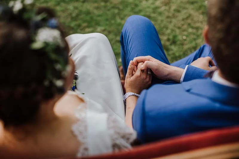 Jeunes mariés se tenant la main, vus de dos, portant une robe blanche et un costume bleu sur l'herbe.
